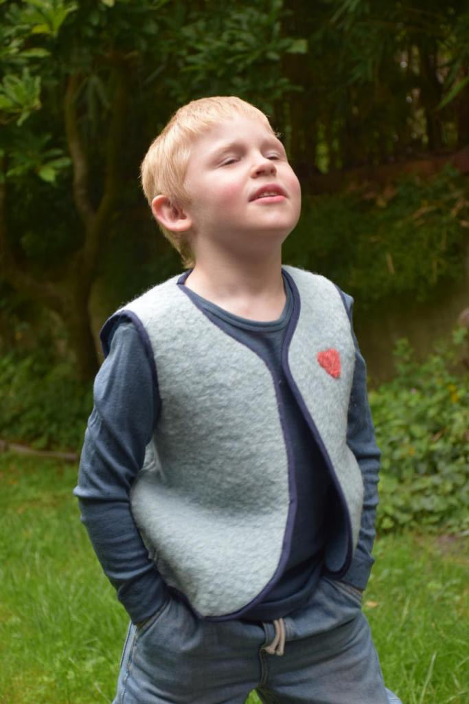 Enfant qui porte un gilet sans manche en laine upcycler de couleur beu avec un coeur rouge sur la poitrine du côté gauche<<<; <vue de profil