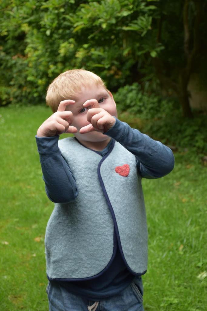 enfant qui porte un gilet sans manche en laine upcycler de couleur beu avec un coeur rouge sur la poitrine du côté gauche. Vue de phase et qui fait un coeur avec ses doigts