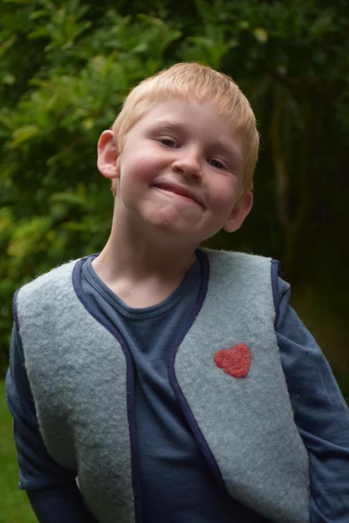 Enfant qui porte un gilet sans manche en laine upcycler de couleur beu avec un coeur rouge sur la poitrine du côté gauche. Vue de face