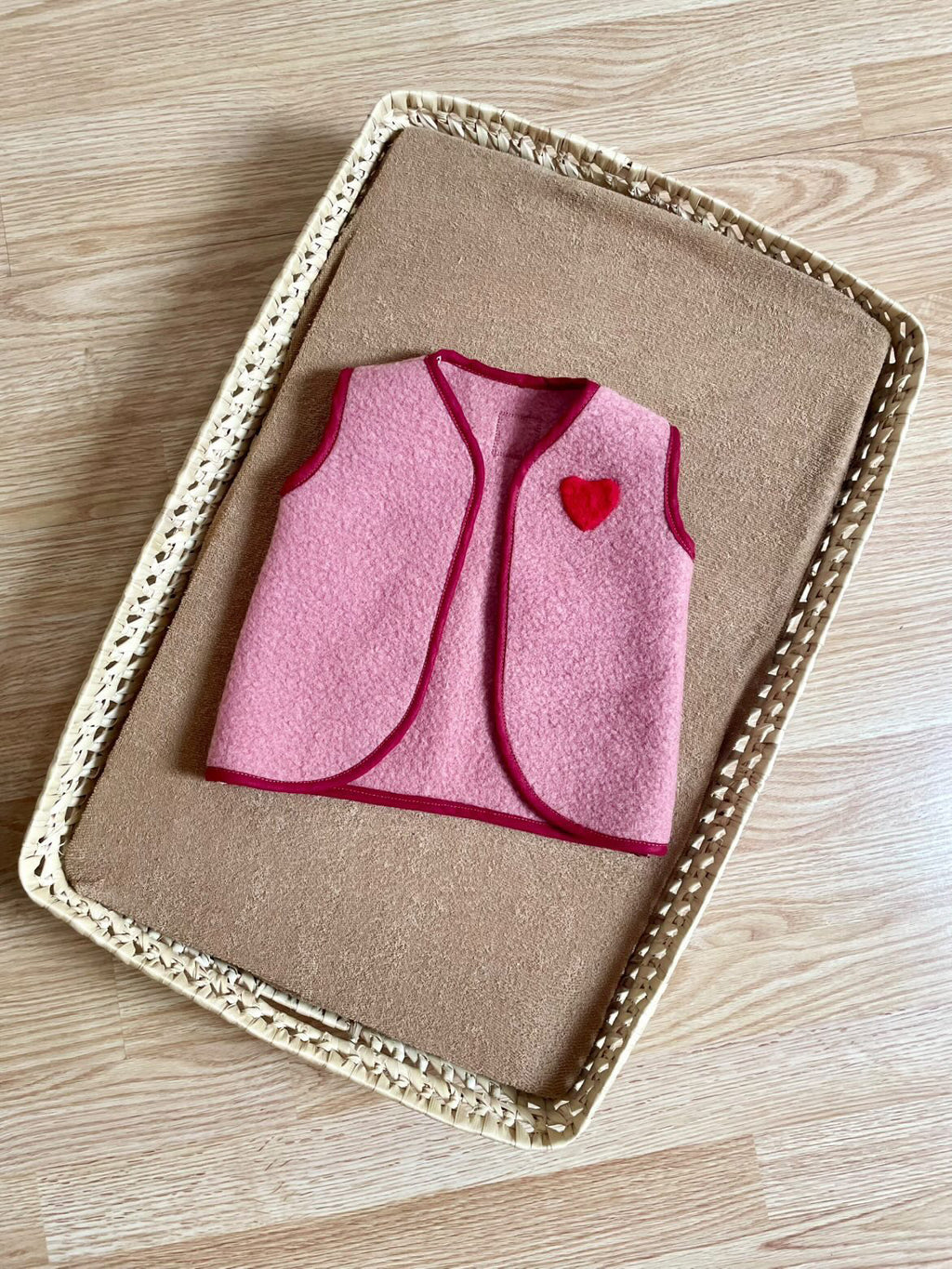 gilet sans manche en laine upcycler de couleur rose avec un coeur rouge sur la poitrine du côté gauche; le gilet est présenté dans un panier en osier