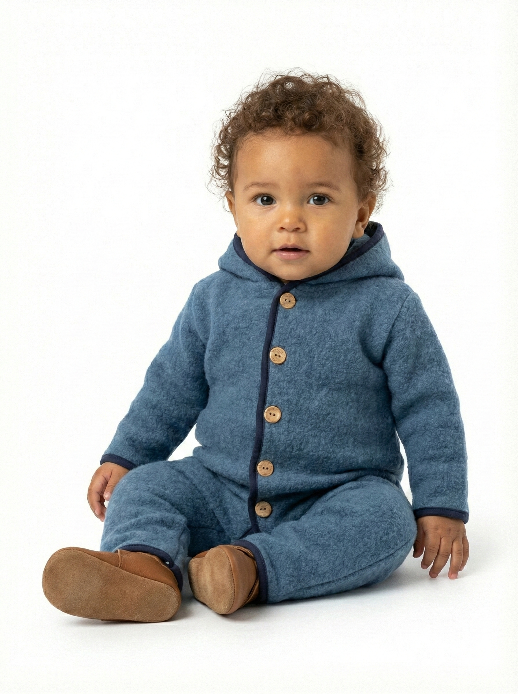 Combinaison pilote à capuche en laine mérinos de couleur bleue, vue de face sur un cintre. Elle est dotée d'une fermeture avant avec des boutons en bois et d'un liseré bleu foncé contrastant le long des bords de la capuche, de la patte de boutonnage, des manches et des pieds intégrés